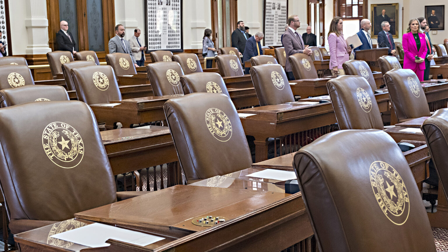 Multiple rows of wood desks and high-backed leather chairs with the Great Seal of the State of Texas embossed on their surface stand empty in a room with wood and glass doors and framed portraits and photos on its walls, with out-of-focus people standing between the desks and chairs on the far side of the space. Multiple rows of wood desks and high-backed leather chairs with the Great Seal of the State of Texas embossed on their surface stand empty in a room with wood and glass doors and framed portraits and photos on its walls, with out-of-focus people standing between the desks and chairs on the far side of the space.