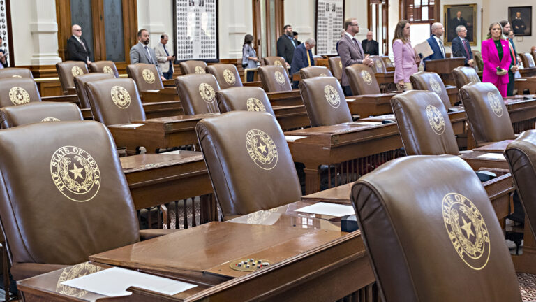 Multiple rows of wood desks and high-backed leather chairs with the Great Seal of the State of Texas embossed on their surface stand empty in a room with wood and glass doors and framed portraits and photos on its walls, with out-of-focus people standing between the desks and chairs on the far side of the space.