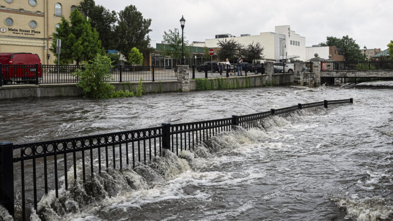 Water flows rapidly through a metal fence along the embankment of a river, with the people standing next to another metal fence atop a taller embankment on the opposite side, with trees, buildings, parked vehicles, streetlights and traffic signs in the background, under a cloudy sky.