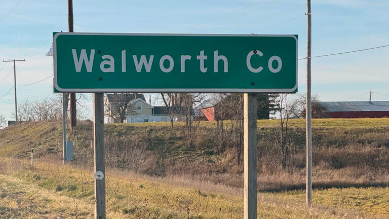 A road sign reading Walworth Co mounted on square wood poles stands along a roadside with an a thinly wooded embankment, utility poles and lines, and farm buildings in the background.