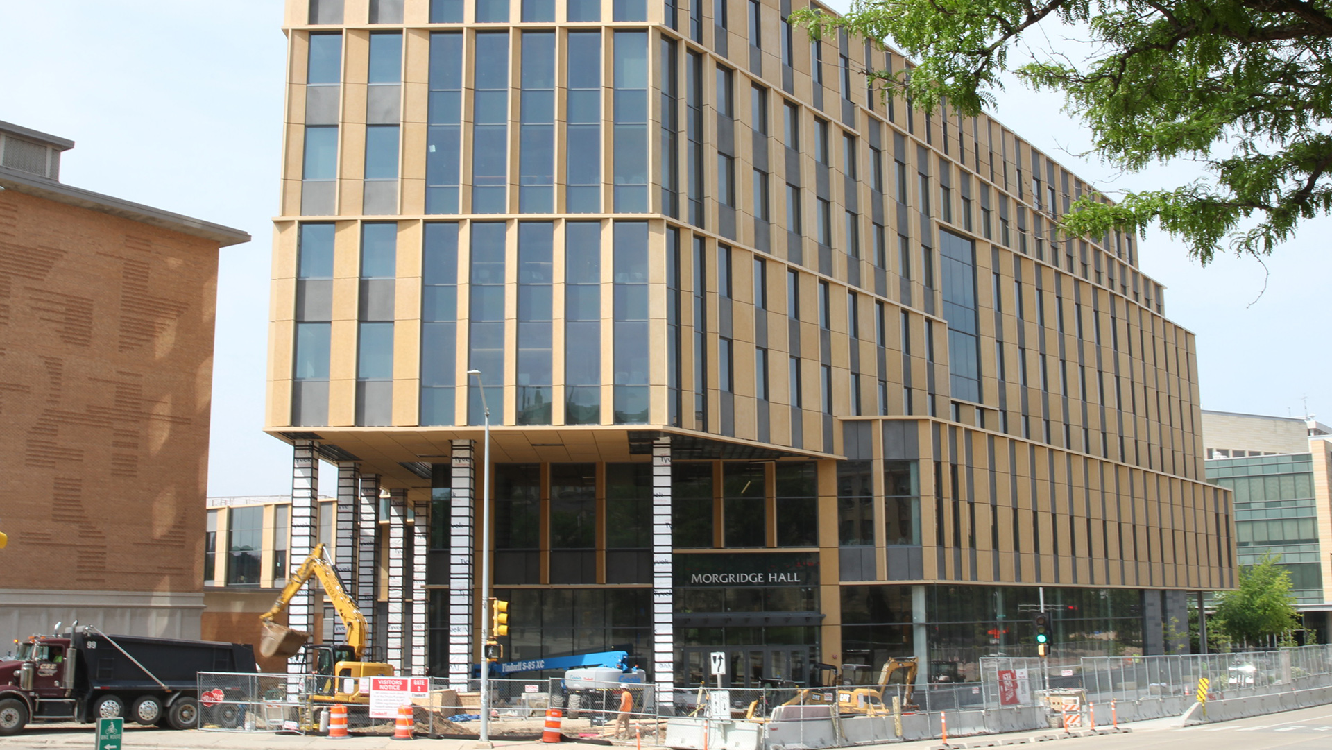 A dump truck and excavator are marked next to other construction equipment, including concrete road barriers, a chain-link fence, traffic barrels and cones, and other items in front of a multi-story stone masonry-and-glass building, with other buildings to the side and in the background, and the leaves of a tree in the foreground.