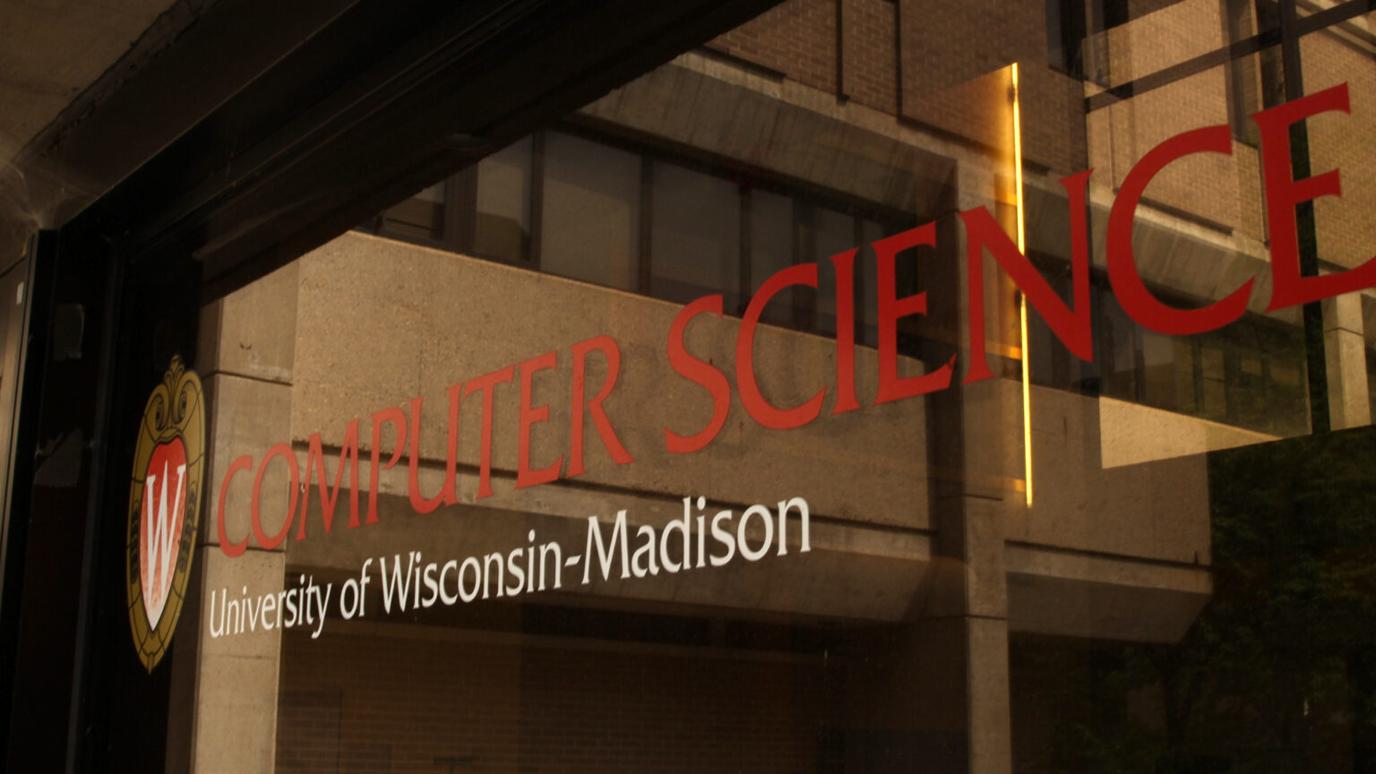 A graphic with UW-Madison's W Crest and the words Computer Science and University of Wisconsin-Madison is attached to a window, with a concrete-and-brick Brutalist-style building visible in its reflection.