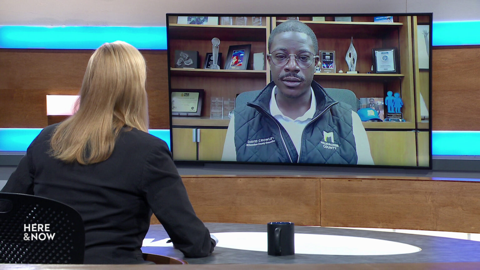Frederica Freyberg sits at a desk on the Here & Now set and faces a video monitor showing an image of David Crowley.