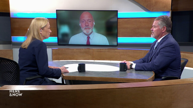 Frederica Freyberg and Bill McCoshen sit on opposite sides of a table on the Here & Now set and face a video monitor showing an image of Scot Ross.