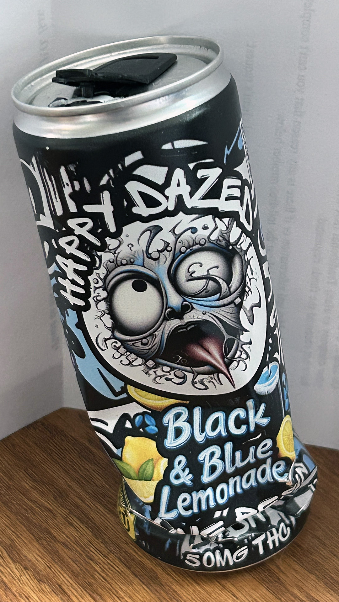 A 16 oz. aluminum can with a plastic tab on its top has a label with an illustration of an abstract face and the words "Happy Dazed," "Black & Blue Lemonade" and "50 mg THC" is propped up in front of a piece of paper on top of a table with a laminate wood surface.