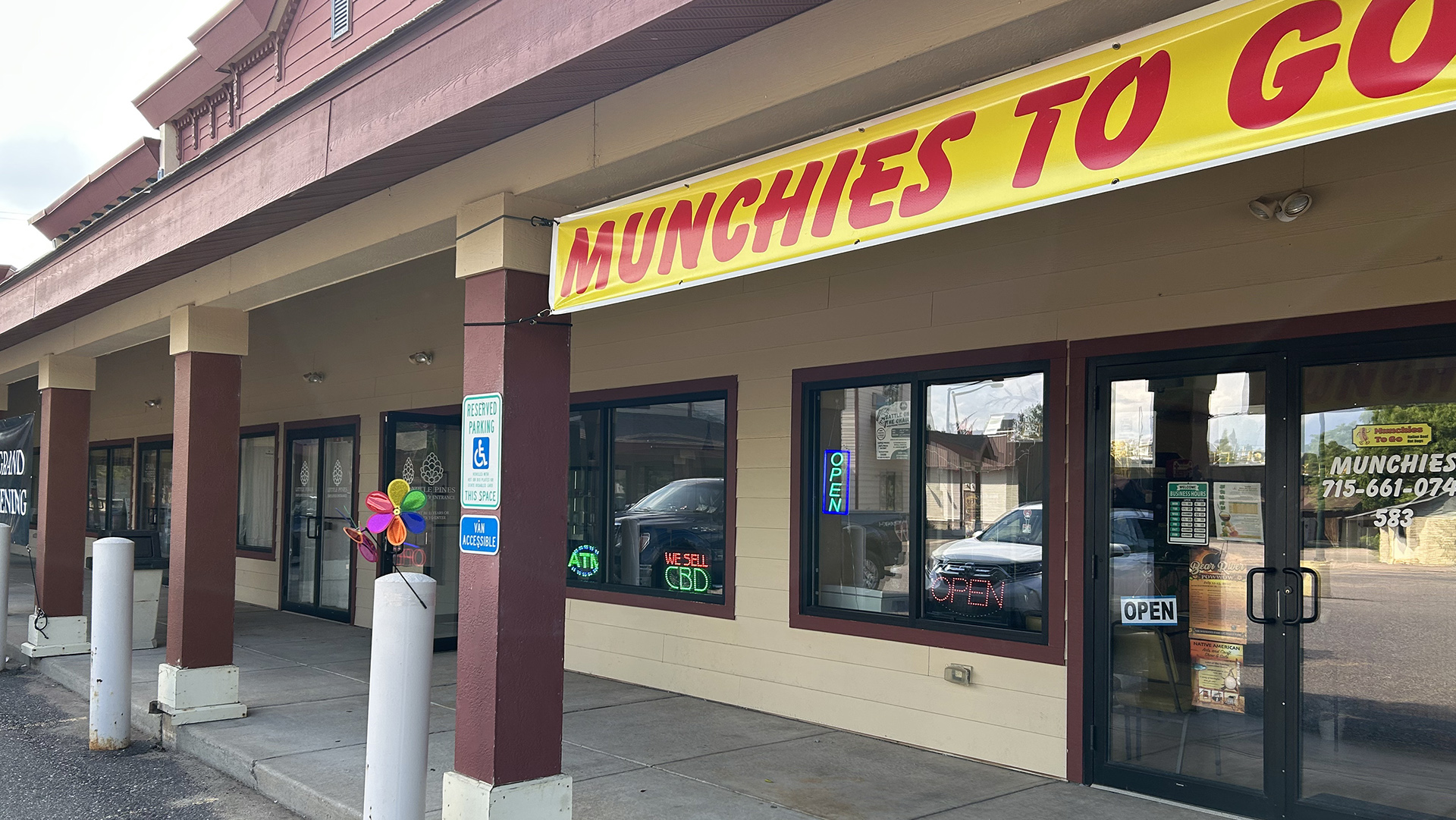 A vinyl sign with the words "Munchies to Go" is connected to the stop of painted square concrete pillars framing a roofed concrete walkway in front of a building with a series of doors and windows, with neon and LED signs reading "Open," "ATM" and "We Sell CBD" in two adjacent windows.