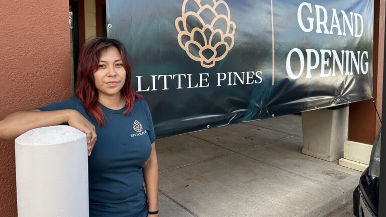 Candace Thompson poses for a photo while standing next to a painted concrete traffic bollard with her right forearm resting on its surface, with a vinyl sign behind her with a logo of a cannabis flower and the words Little Pines and Grand Opening connected to painted concrete square pillars framing a concrete sidewalk in front of a building.
