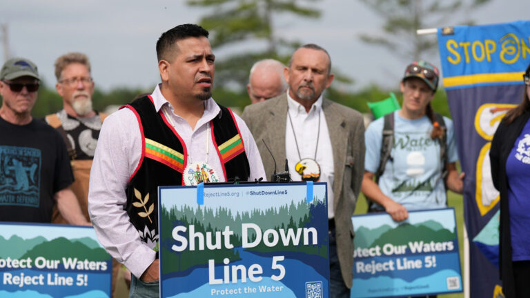 A man wearing a tribal regalia vest and medallion speaks while standing behind a podium with a political yard sign with a graphic of trees and water and the words Shut Down Line 5 and Protect the Water affixed to its front, with other people standing behind him, including several holding the same sign.