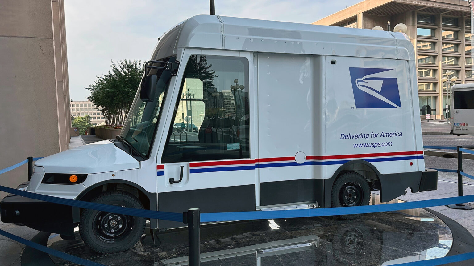 A Next Generation Delivery Vehicle with the logo of the U.S. Postal Service, the words Delivering for America and the URL www.usps.com on its side is parked on a round platform and surrounded by a retractable belt barrier next to a stone masonry building, with trees in concrete planters and other buildings in the background.
