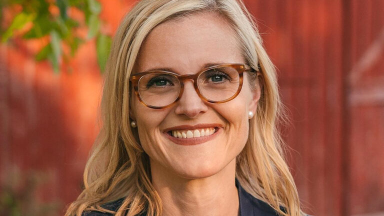 Sarah Godlewski poses for a portrait with out-of-focus leaves and a painted wood wall in the background.
