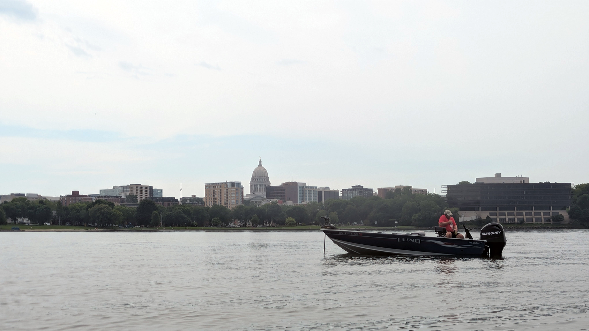 An angler sits near the stern of an aluminum fishing boat with an outboard motor in a calm lake, with the skyline of a city with a domed building at center and a park with lawn and trees on the shoreline in the background, under a cloudy and hazy sky.