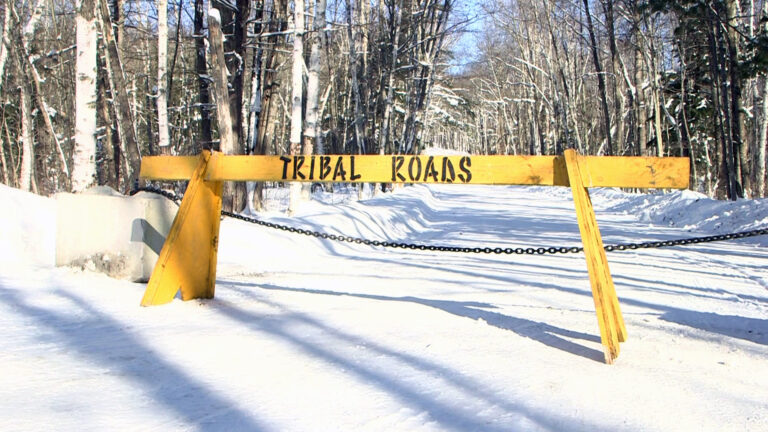 A wood road barrier with tribal roads painted on it and chain connected to a concrete block spans a snow-covered road with trees on either side.