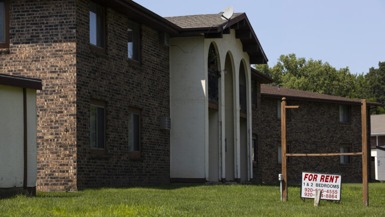 A yard sign with the words For Rent and 1 & 2 Bedrooms along with two phone numbers stands in a lawn under an empty wood frame for a sign, next to a two-story brick and stucco building with three two-story arches framing patios and decks, with the tops of trees behind the building in the background.