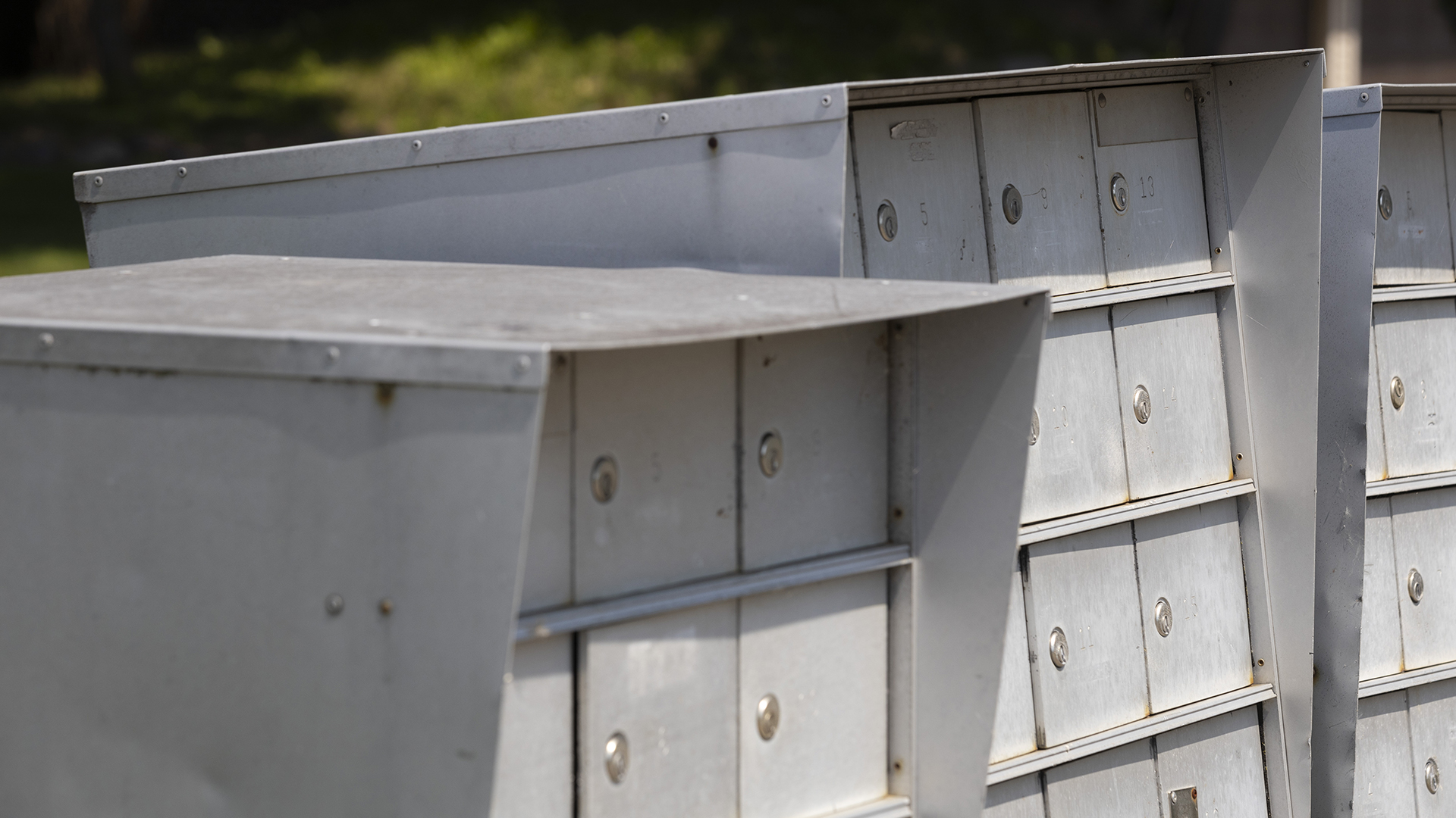 Sunlight illuminate and shadows obscure different areas of multiple aluminum cluster mailboxes with rows of doors with keyholes.
