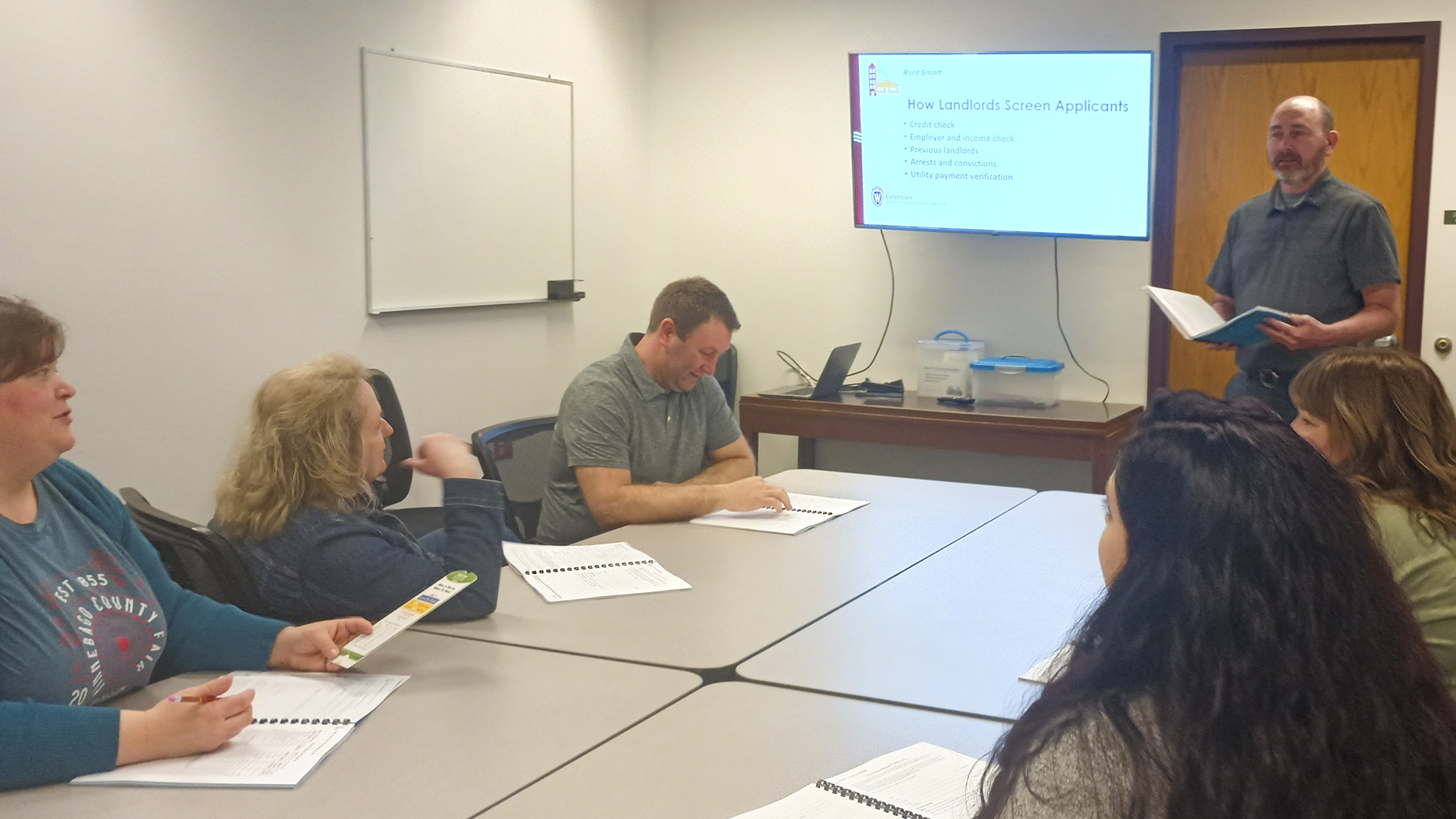 Todd Wenzel stands with an open book in his hands and faces four connected tables with five people sitting on either side with open spiral-bound books in front of them, in a room with a closed wood door, a table with an open laptop computer and two closed plastic storage bins on its surface, a monitor with the words "How Landlords Screen Applicants" on its screen, and a clean whiteboard.