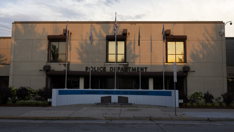 The U.S. and other flats are displayed atop five flagpoles situated in a row on a concrete platform along a sidewalk and road and in front of a multi-story masonry building with sunlight reflecting off the windows on its second story, with a security camera mounted on one corner of its roof.