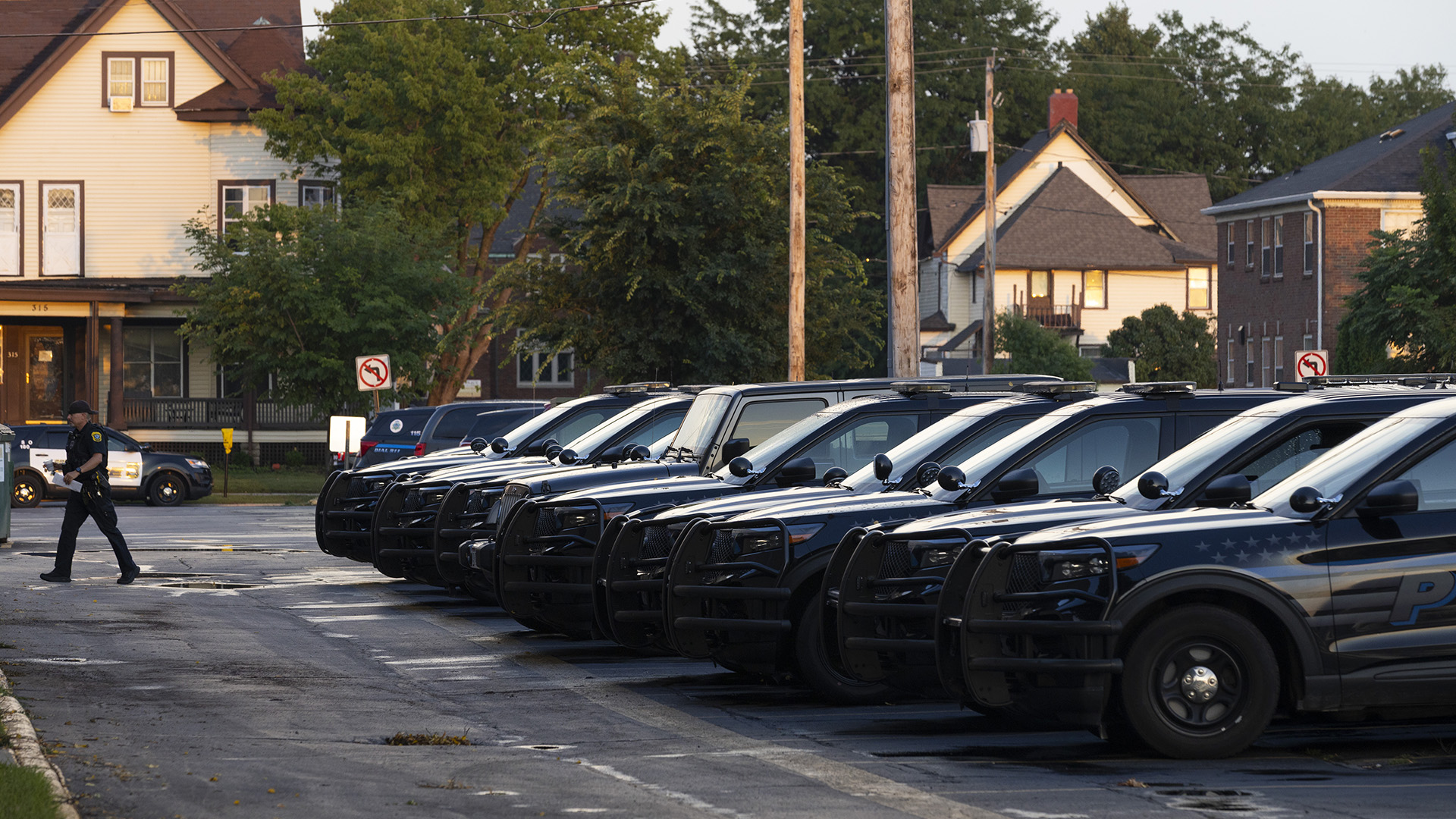 A police officer walks across an asphalt parking lot with puddles of water on its surface away from a row of multiple parked police vehicles, with other police vehicles parked along a road in the background in front of multiple houses, trees and utility polls.