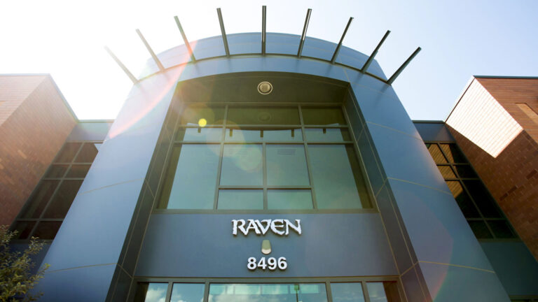 A lens flare centers on sunlight shining above a multistory building with brick and metal siding, with large plate-glass windows above a sign reading Raven and an address number of 8496 marking a central entrance topped by a crown of metal rods emanating from the wall near the roof.