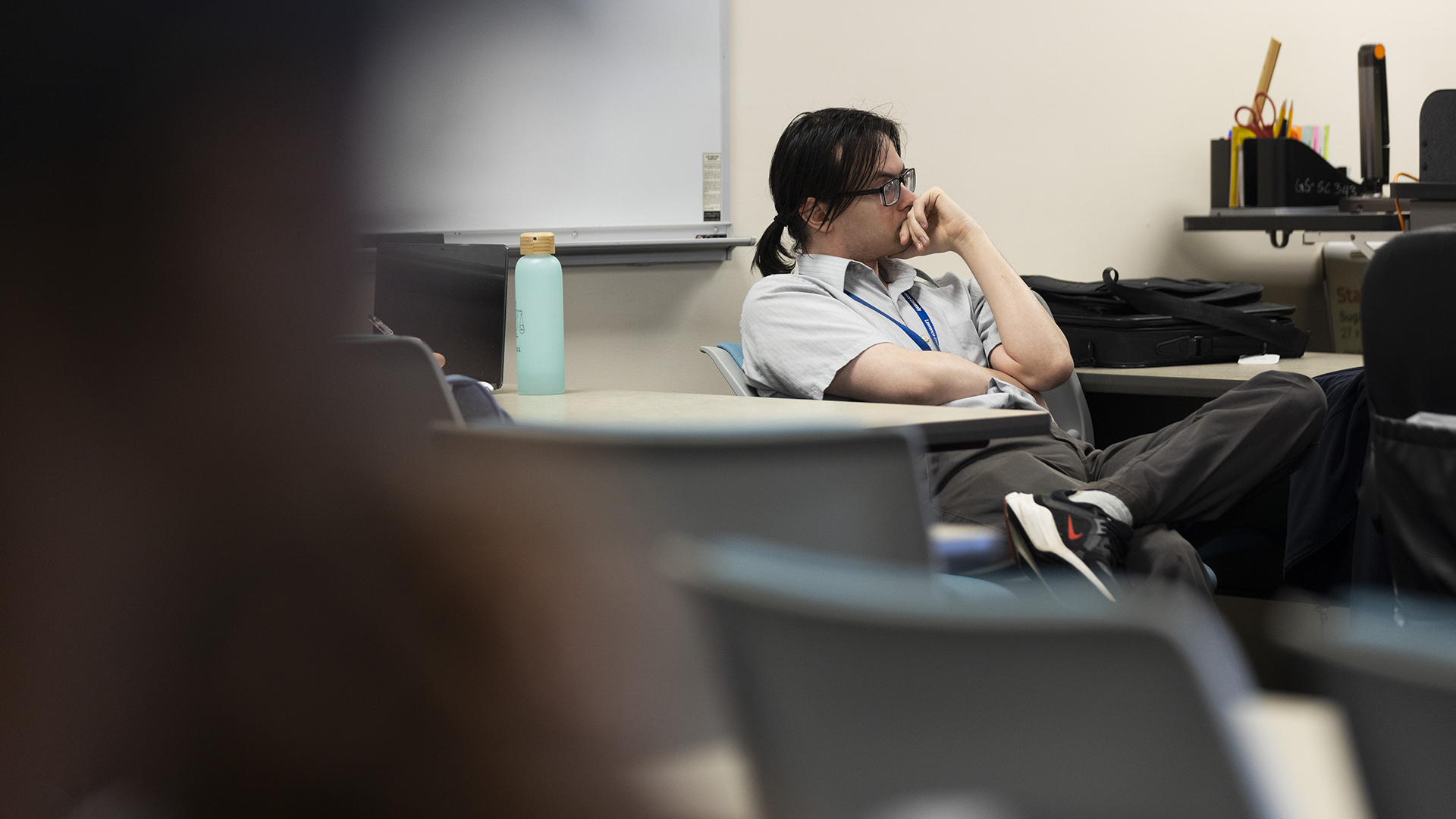 Nick Bengry sits with crossed legs on a reclining office chair in a room with multiple rows of tables, a desk with a ruler, scissors, pencils, highlighters and other items on its surface, and a whiteboard on a wall, with an out-of-focus person seated in the foreground.