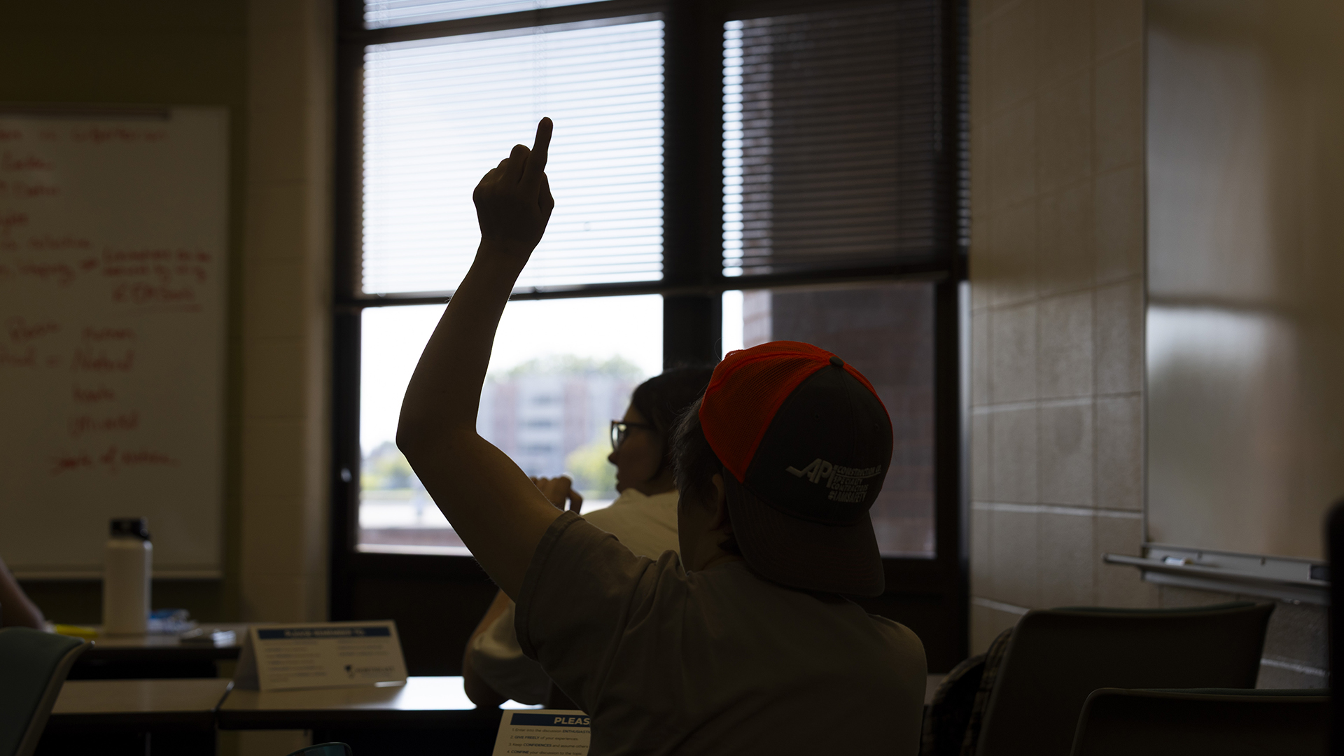 A seated person in silhouette raises their left hand in the air while seated behind another person in a room with a window with half-drawn blinds and an out-of-focus building visible outside.