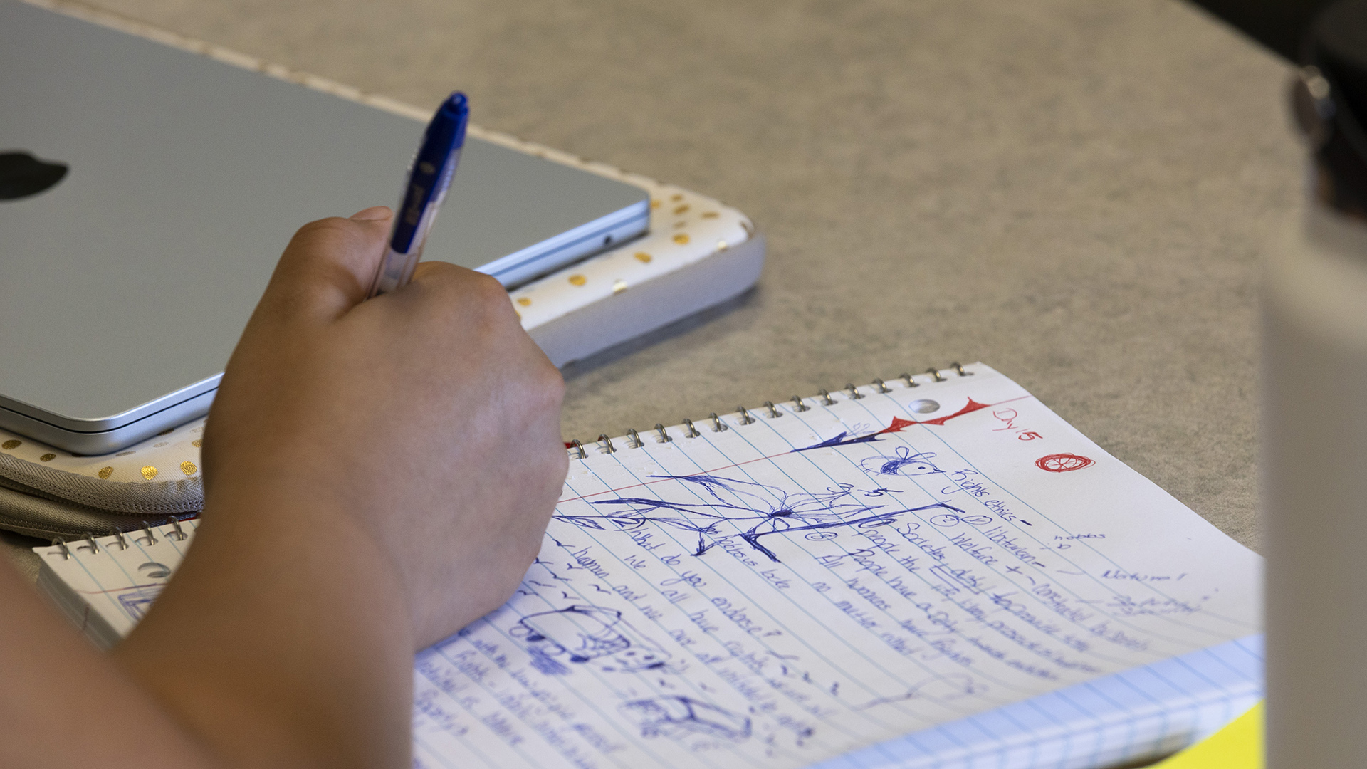 A right hand holding a pen writes on a notebook with lined paper with notes and doodles on the top page that is sitting on top of a table next to a closed laptop computer resting on top of a laptop sleeve.
