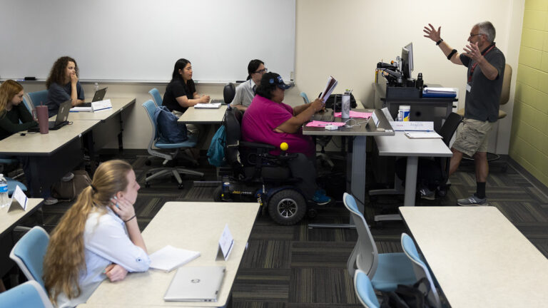 Patrick Parise gestures with both hand in the air while standing in front of several rows of tables with students sitting in rolling chairs behind them, in a room with a low pile carpet, a painted concrete block wall and a large whiteboard.