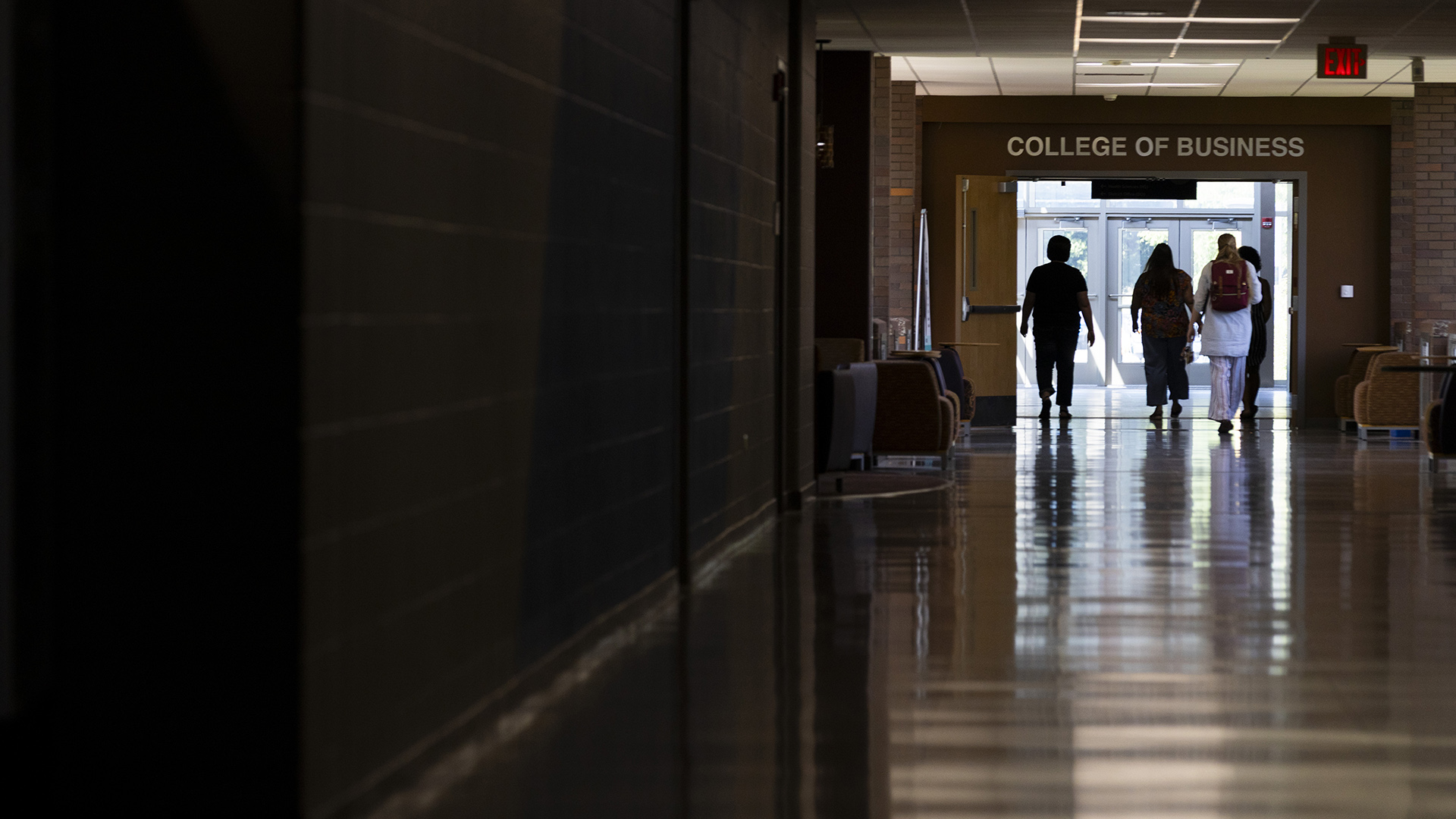 Four students walk down a hallway through open double doors with push handles under a sign reading "College of Business" toward multiple metal and glass doors to the outside.