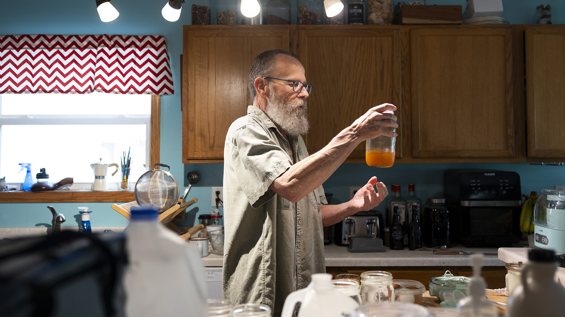 Earl Lowrie stands in a kitchen and holds a mason jar about one-third filled with a beverage, with a window with a valance, a row of cabinets with different items on the top of each and a countertop with different appliances and bottles on its surface, with an out-of-focus assortment of plastic and glass containers in the foreground.