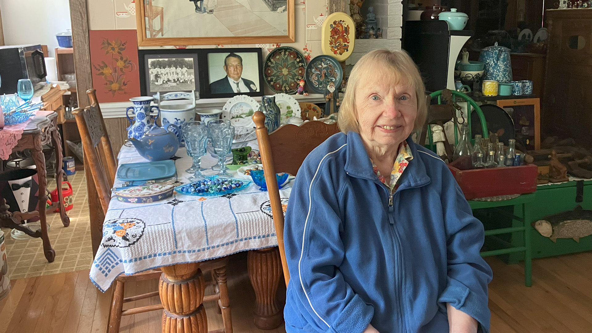 Pam Goodlet poses for a portrait while sitting on a high-backed wood chair, with wood tables, chairs and shelves in the background covered with plates, glasses, bottles and other items, with photos and paintings mounted on a wall in the background.