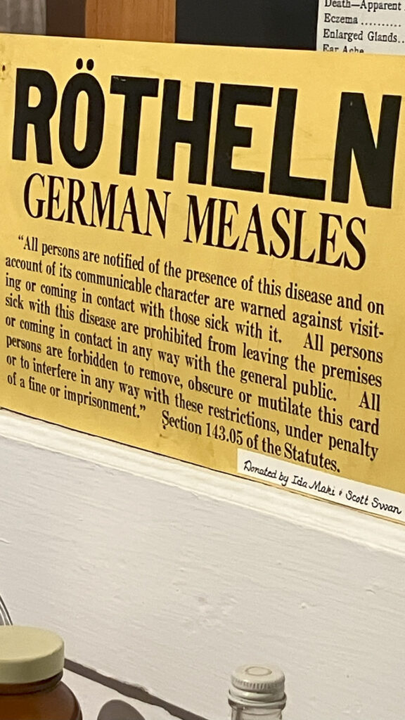 A printed sign with the words "Rötheln" and "German Measles" in large type followed by a quotation of "Section 143.05 of the Statutes" is propped up on the edge of a windowsill above multiple bottles,