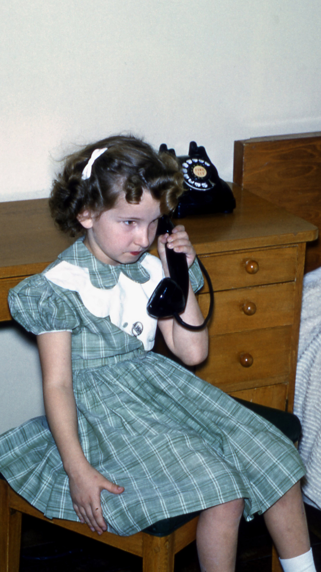 Dorothy Thompson holds the receiver of a rotary dial telephone in her left hand while siting on a wood bench with a padded top next to a wood desk and bed.