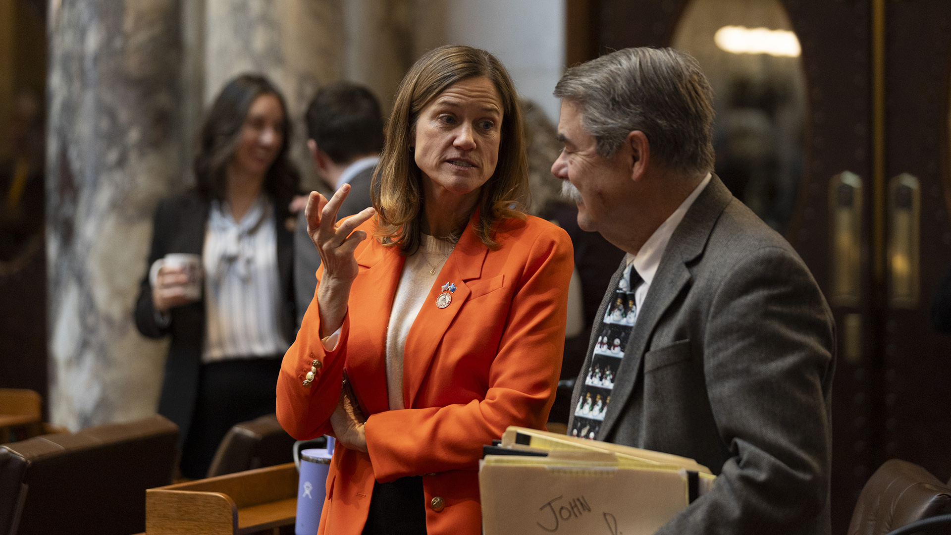 Deb Andraca gestures with her right hand while speaking and standing next to Joe Sheehan, with other out-of-focus people standing in the background of a room with marble pillars and double doors with oval windows.