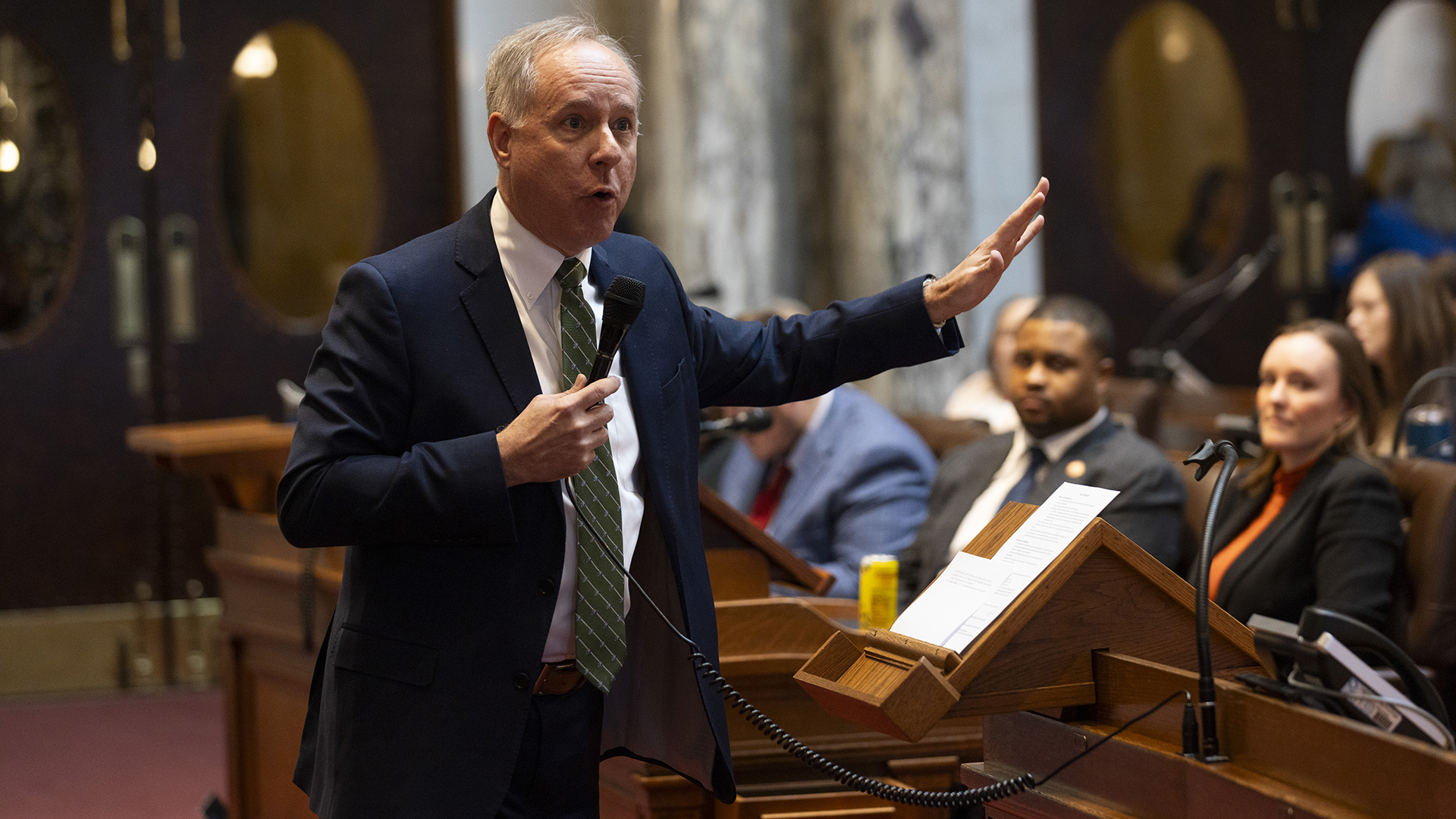 Robin Vos gestures with his left hand and speaks into a corded microphone in his right hand, while standing in front of a wood podium with printed papers on its surface, while standing and facing out-of-focus people seated in rows of desks, in a room with marble pillars and double doors with oval windows.