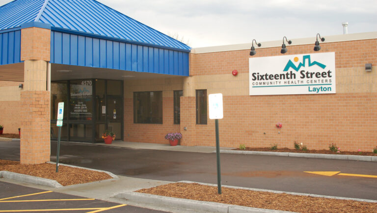 A sign with a logo of a skyline and the words Sixteenth Street Community Health Centers and Layton is mounted to the size of a brick-sided building under three spotlights mounted to the edge of a room, with a pyramidal metal-sided awning supported by brick-sided pillars covering a glass-walled entrance next to a paved driveway with a direction arrow and mulch-covered berm with parking signs mounted to perforated metal poles.