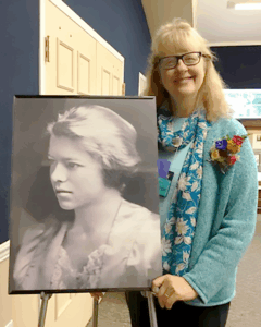 Smiling woman with glasses stands indoors beside a black-and-white portrait of poet Lorine Niedecker displayed on an easel. She wears a turquoise sweater with a floral scarf and colorful flower pin.