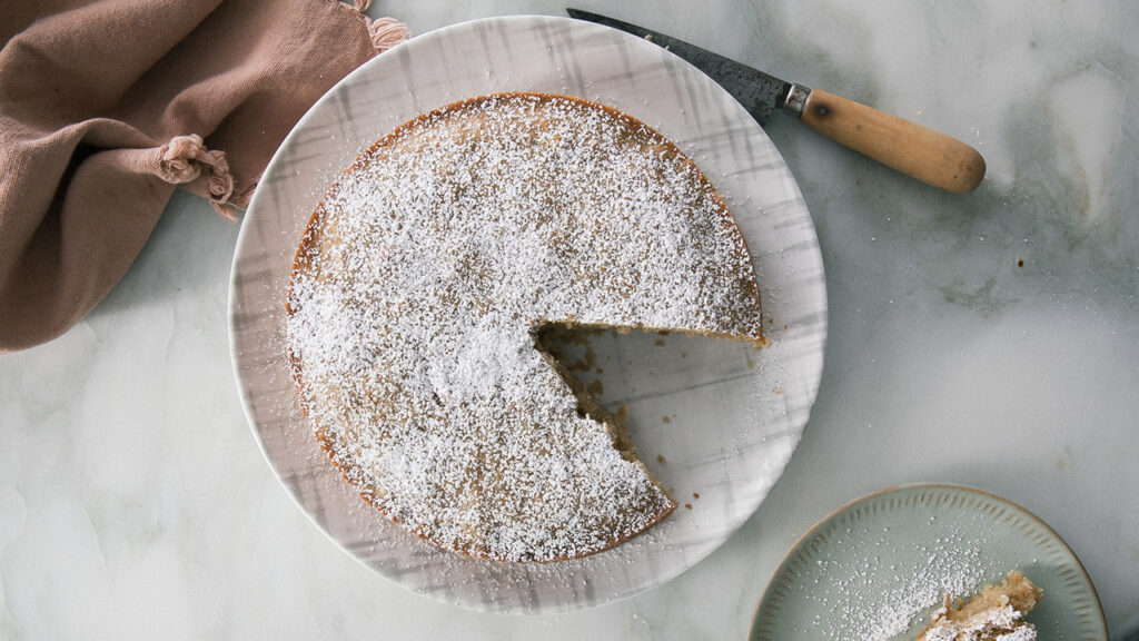 An apple and olive oil cake adorned with powdered sugar, with a missing a piece.