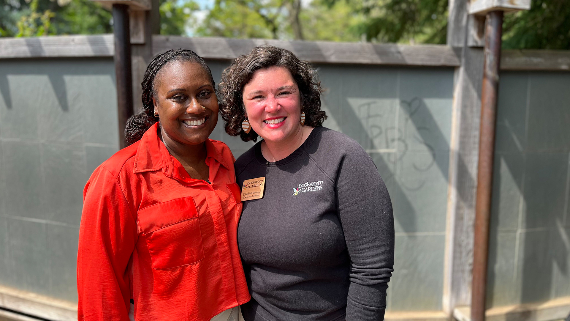 Two women stand side by side - 'Wisconsin Life' host Angela Fitzgerald on the left and Bookworm Gardens executive director Elizabeth Wieland - on the right in front of a wooden fence, smiling.