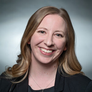 Professional headshot of a woman with long wavy hair, smiling, wearing a blazer and pendant necklace, in front of a gradient background.