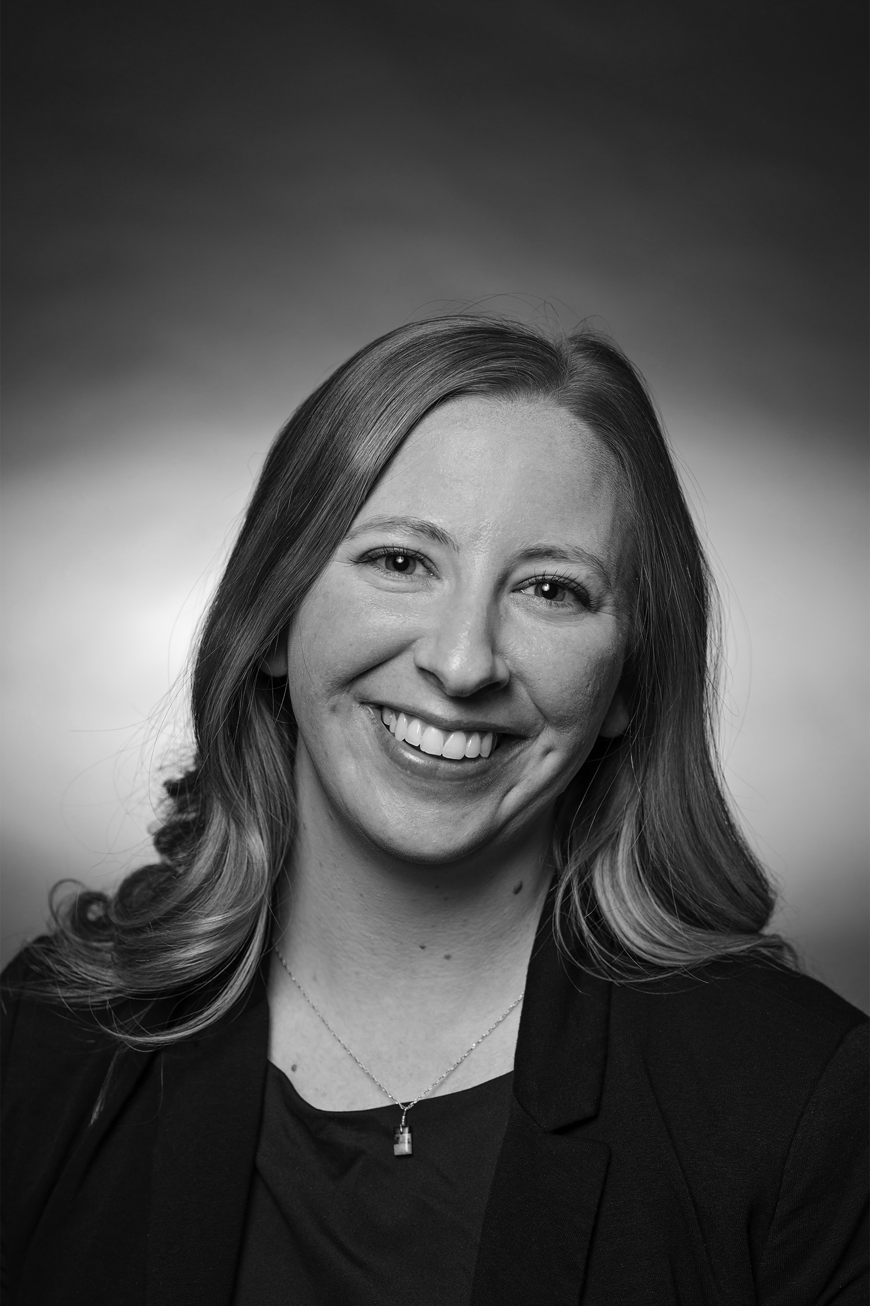 Professional headshot of a woman with long wavy hair, smiling, wearing a blazer and pendant necklace, in front of a gradient background.