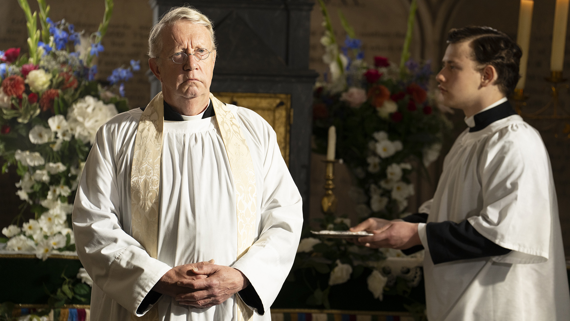 A priest in white robes and a gold stole stands at the front of a church staring at the churchgoers with a serious expression.