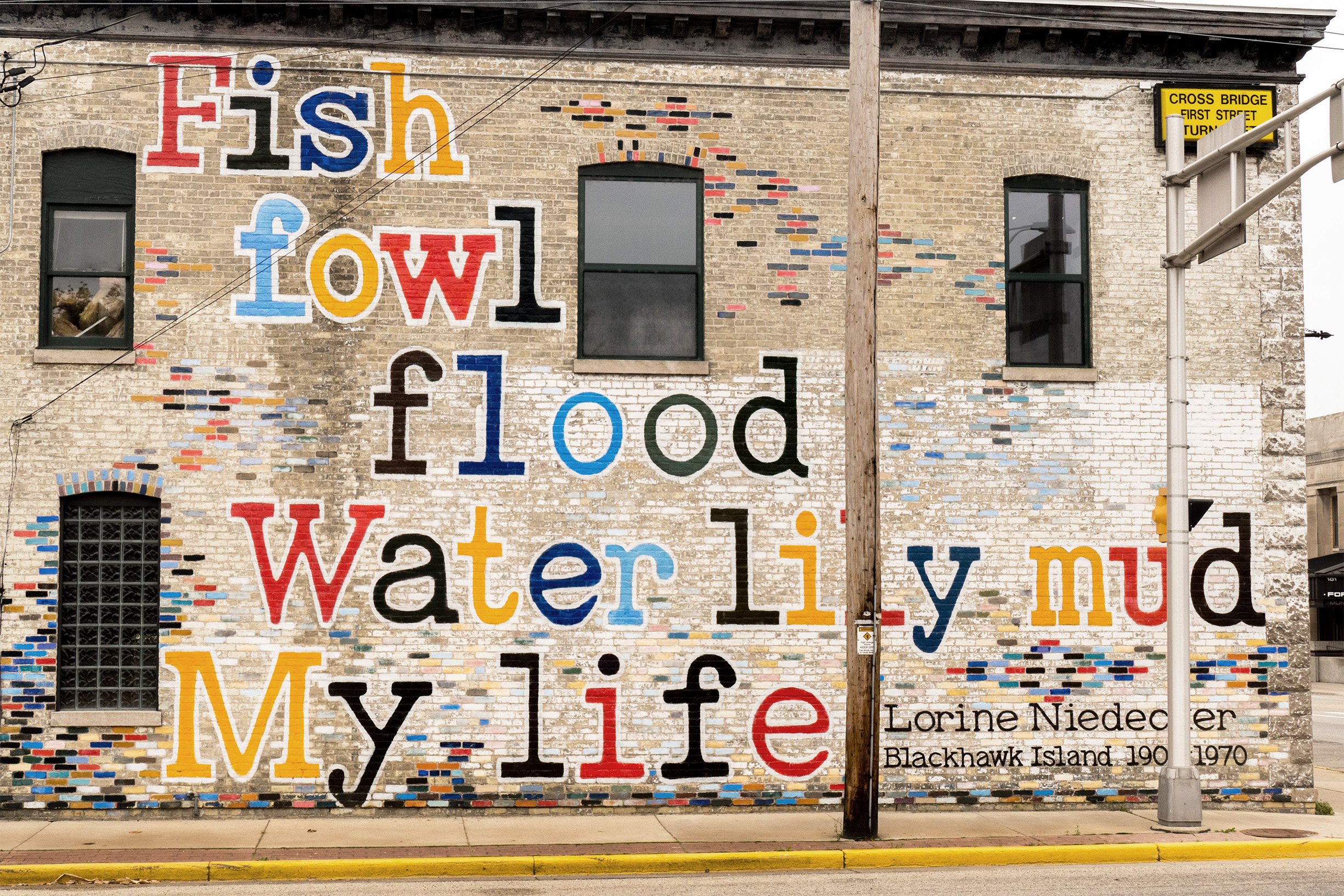 Mural on a brick building with the words “Fish/ fowl/ flood/ Water lily mud/ My life” painted in large colorful letters. At the bottom, text reads “Lorine Niedecker, Blackhawk Island 1903–1970.”