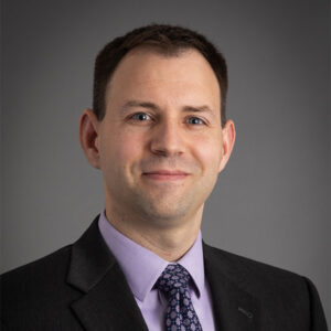 Professional headshot of a man in a dark suit and patterned tie, smiling slightly, in front of a plain background.
