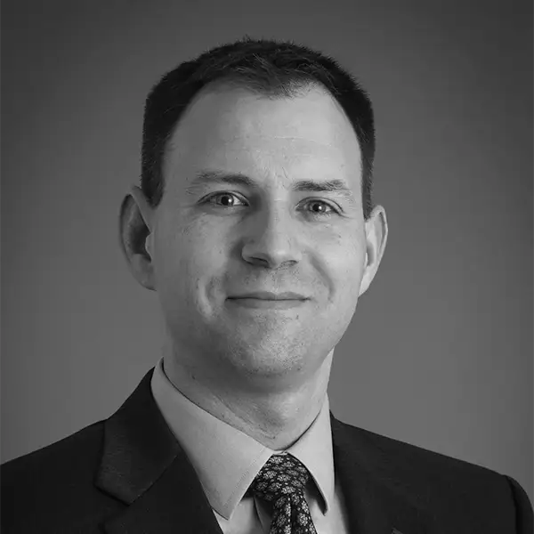 Black-and-white portrait of a man in a suit and patterned tie, smiling slightly against a neutral background.