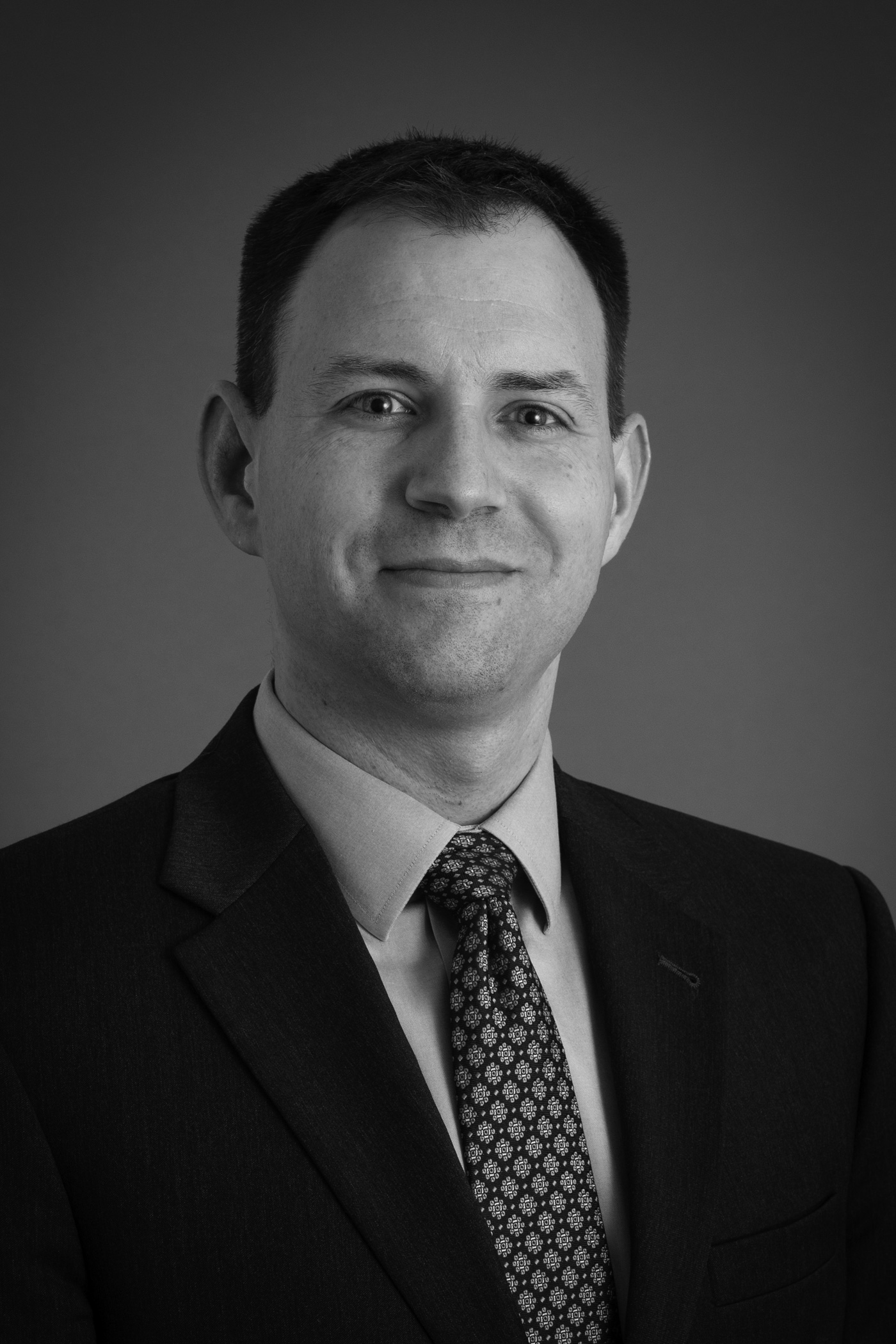 Professional headshot of a man in a dark suit and patterned tie, smiling slightly, in front of a plain background.