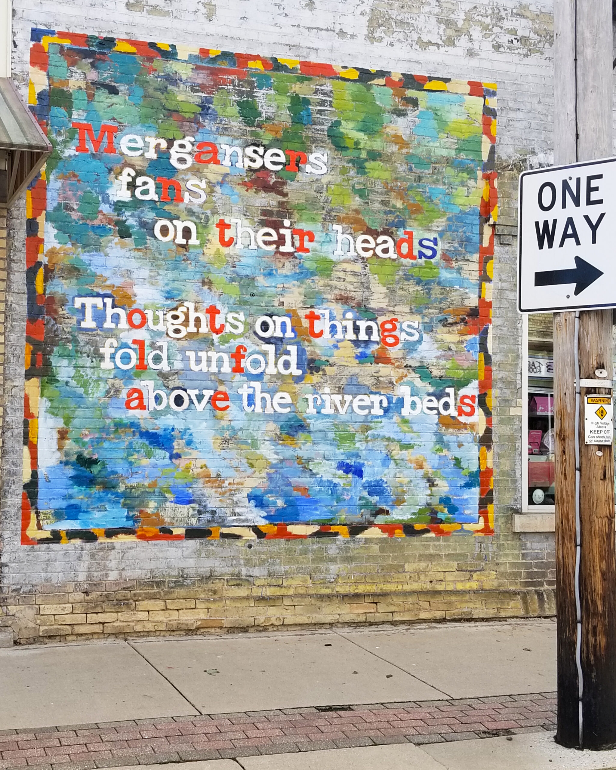 A colorful mural painted on a brick wall shows abstract brushstrokes in greens, blues, and yellows, with words in bold red, white, blue, and black letters reading: “Mergansers/ fans/ on their heads/ Thoughts on things/ fold unfold/ above the river beds.”