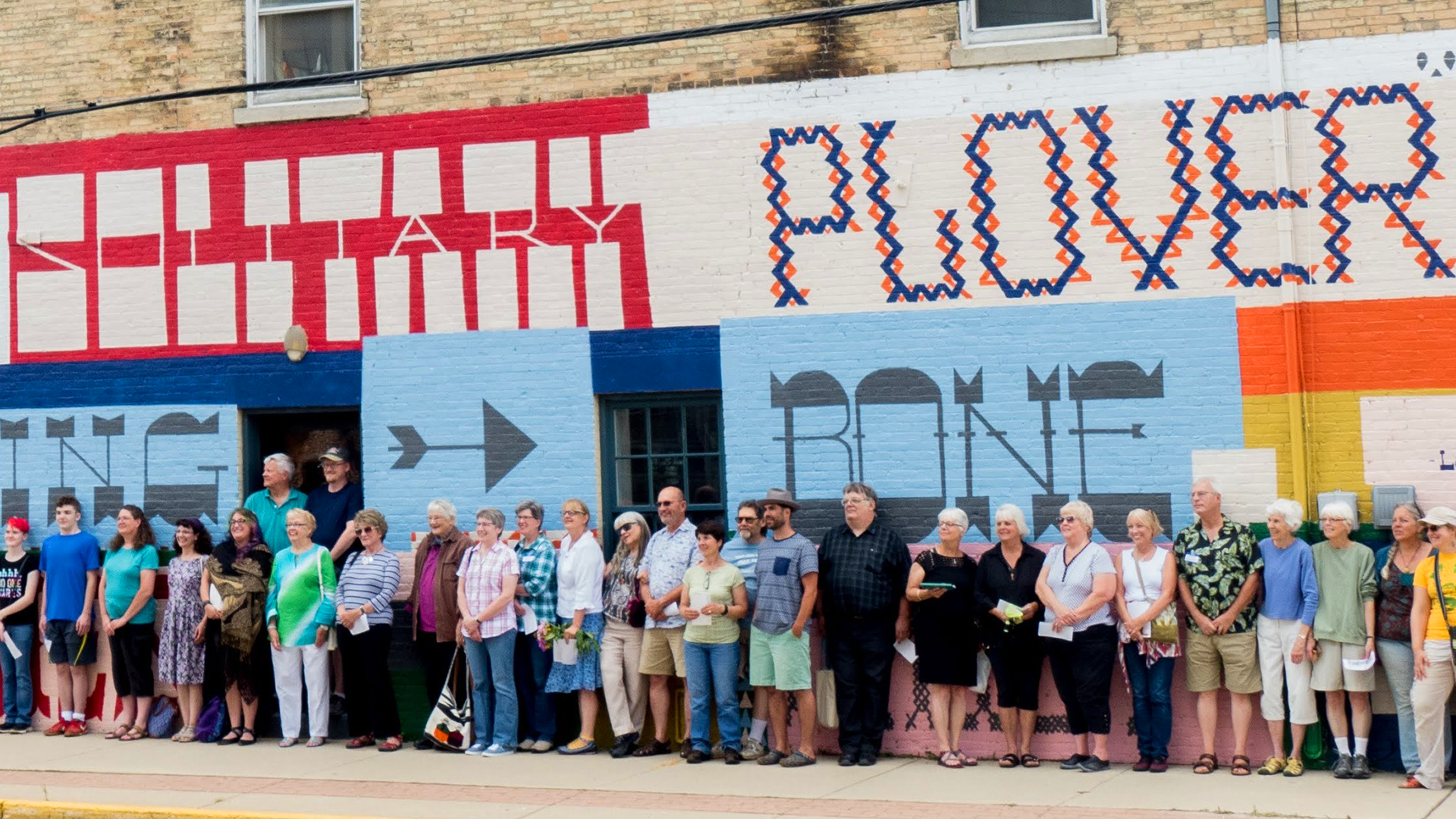 A large group of people stand in a line on the sidewalk in front of a colorful mural painted on a brick wall. The mural features bold, geometric lettering in red, blue, black, and orange. Part of the text reads “SOLITARY PLOVER,” with “SOLITARY” in blocky red-and-white letters and “PLOVER” in a patterned blue-and-orange style. Additional words in black and gray, along with arrows and decorative elements, fill the mural’s background. The group consists of people of various ages, casually dressed, some holding papers or bags, appearing gathered for a community or celebratory event.