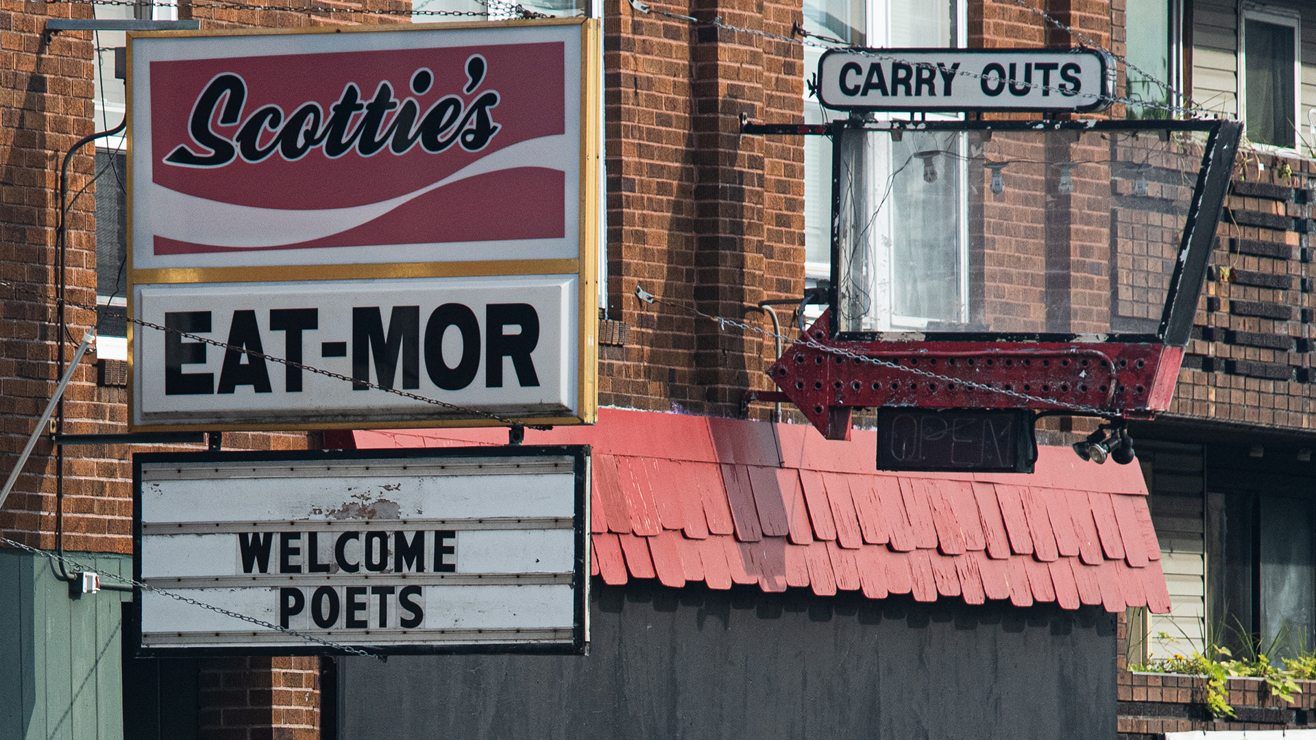 Close-up of signs outside a brick building. A red-and-white sign reads “Scottie’s EAT-MOR,” and a changeable letter board below says “WELCOME POETS.” To the right, an old arrow sign points toward a “CARRY OUTS” sign.