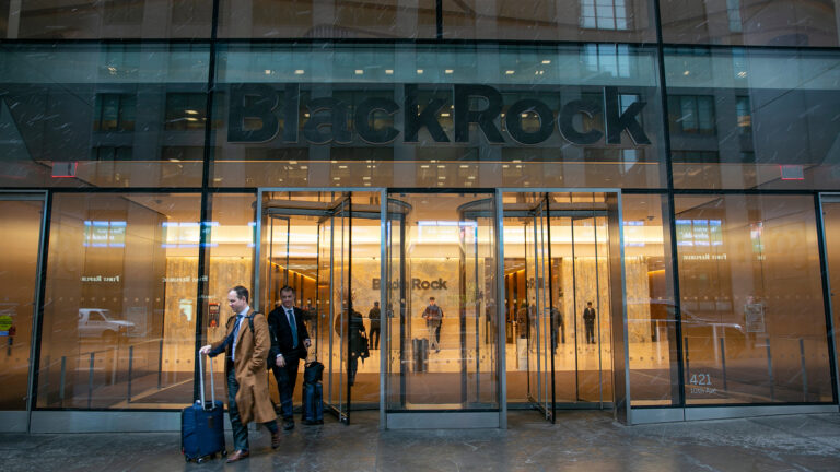 Two men holding the handles of rolling carry-on luggage pass through a revolving door in the glass-walled entrance of a building with a letter sign on the exterior reading BlackRock and with more people standing inside an illuminated lobby with a smaller letter sign reading BlackRock and hallways leading to rows of elevators.
