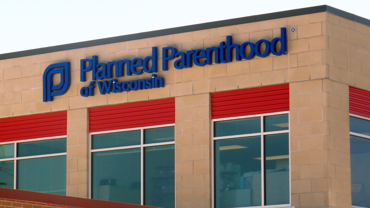 A letter sign with the words Planned Parenthood of Wisconsin and a logo is affixed to the side of a brick building with multiple windows.