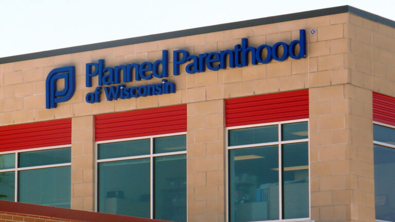 A letter sign with the words Planned Parenthood of Wisconsin and a logo is affixed to the side of a brick building with multiple windows.